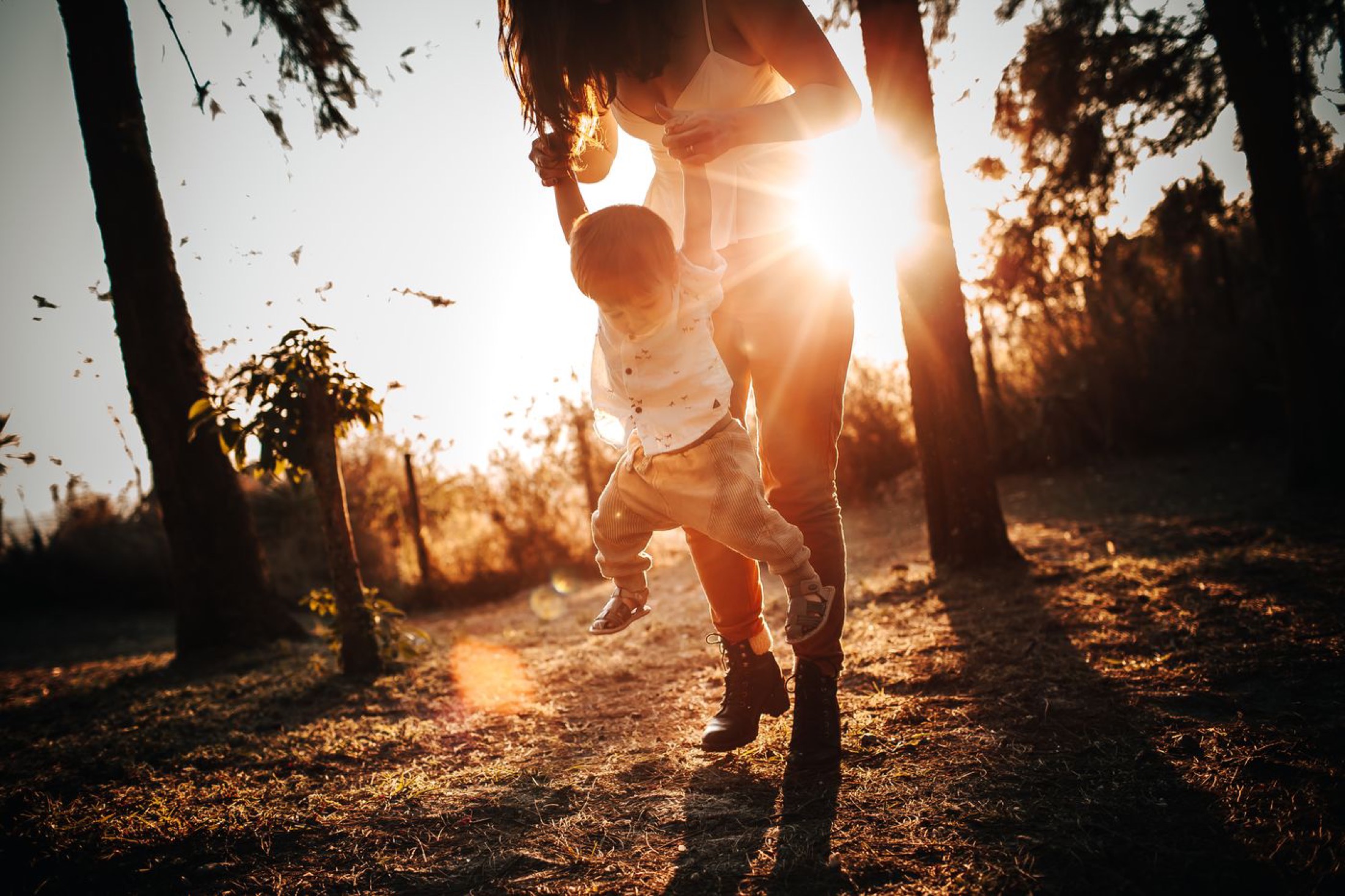 A mother swinging her small child by the hands in a park at sunset.